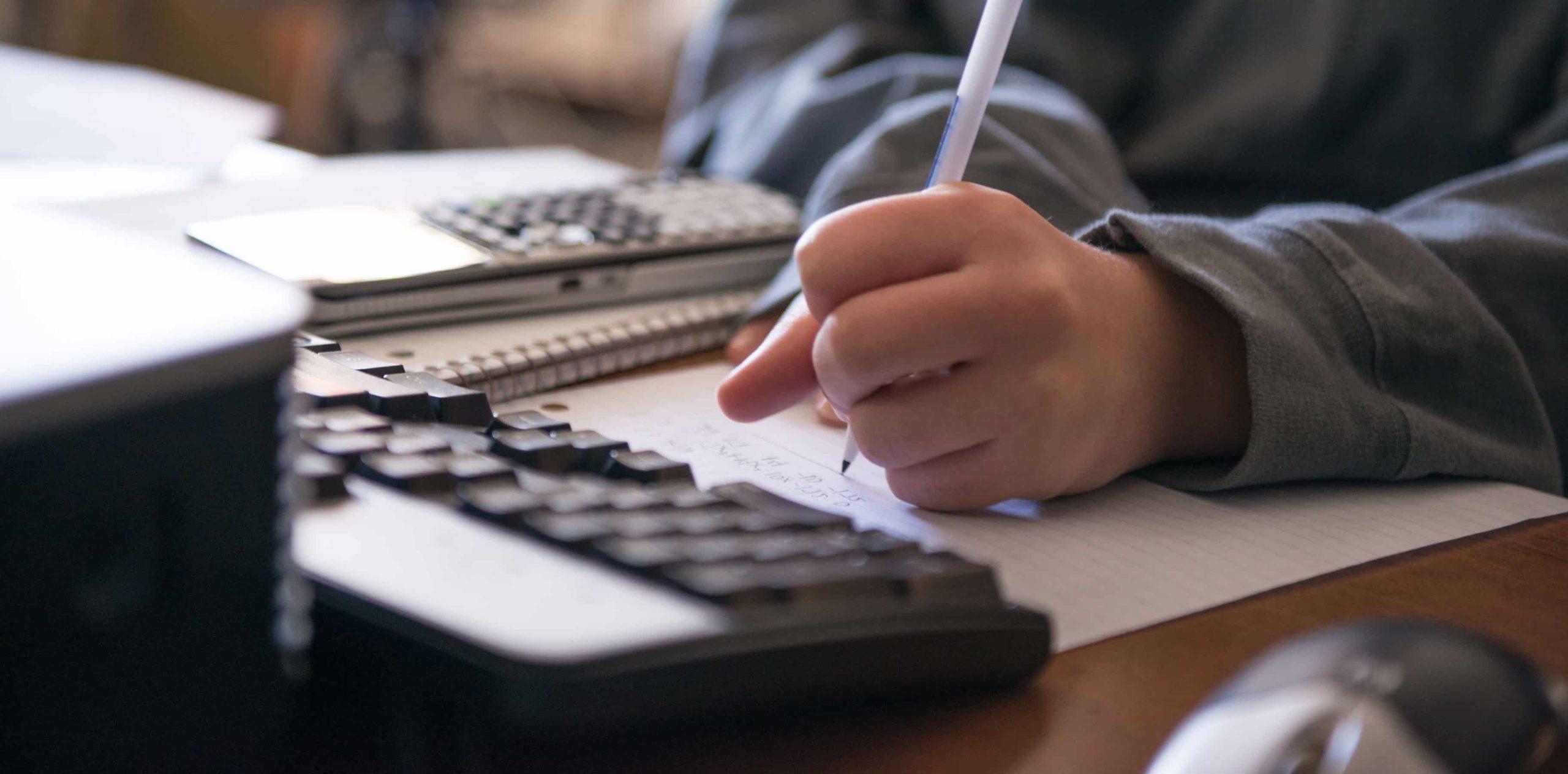 Someone appearing to do a math problem on a sheet of paper with a computer in front of them and a calculator on the side.