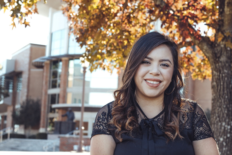 A young woman smiling with brown curled hair. She wearing a black short sleeve blouse. She is standing by a tree with autumn colored leaves. In the background there is a building with a lot of windows.