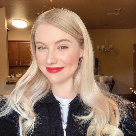 Portrait of a smiling woman with long, wavy platinum blonde hair, wearing a black zip-up top. She has bright red lipstick and is posing indoors, with soft lighting illuminating her face. The background includes part of a living space, with wooden cabinets and a light fixture visible.