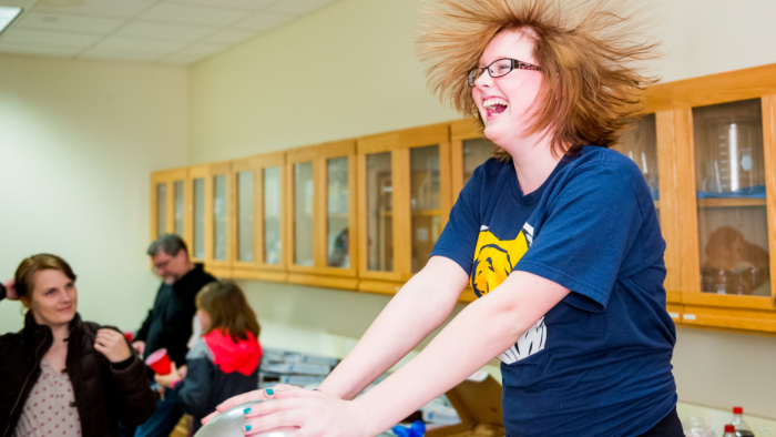 A student touches a van de graaff generator, resulting in their long, red hair standing on end.